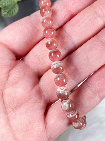 A close-up shot captures a hand holding a string of The Crystalary's Rhodochrosite Crystal Bracelet, featuring polished, round pink beads with a slightly translucent quality. The bracelet highlights a natural, marbled pattern. In the background, a potted plant with green leaves subtly enhances the serene ambiance that symbolizes self-love.