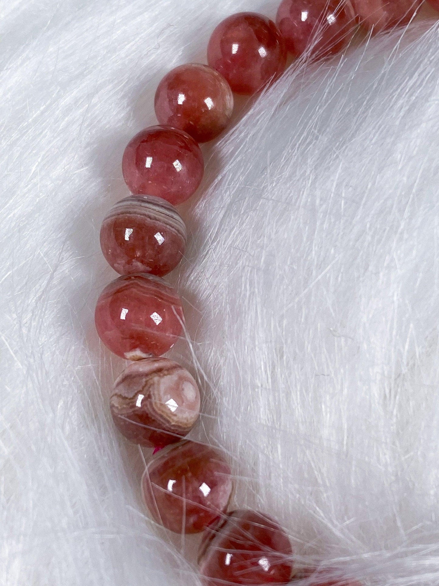 A Rhodochrosite Crystal Bracelet from The Crystalary is arranged in a semi-circle on a soft white fur background. The beads exhibit an array of patterns and hues, creating texture and color contrast against the fur, evoking the allure of gemstone bracelets that signify self-love.