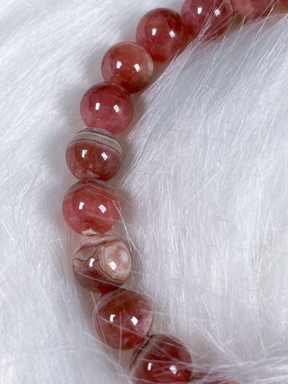 A Rhodochrosite Crystal Bracelet from The Crystalary is arranged in a semi-circle on a soft white fur background. The beads exhibit an array of patterns and hues, creating texture and color contrast against the fur, evoking the allure of gemstone bracelets that signify self-love.