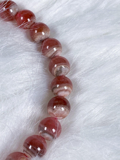 A close-up of The Crystalary's Rhodochrosite Crystal Bracelet, featuring polished, round, reddish-brown and white stones. Known for its self-love aura similar to other Rhodochrosite crystal bracelets, it is elegantly resting on a soft, white, fur-like surface. The beads showcase charming variations in color and patterns.