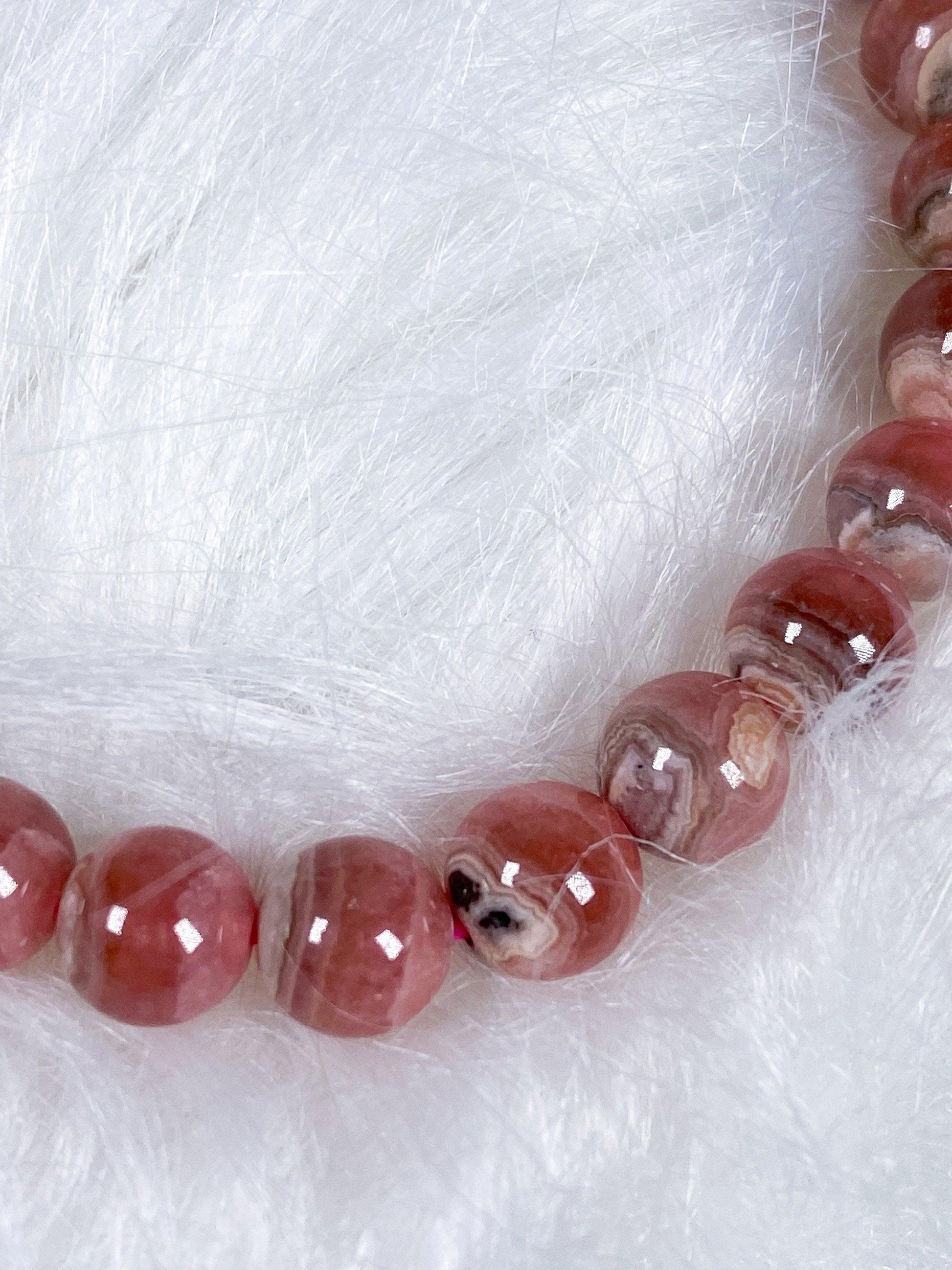 A close-up view of The Crystalary's Rhodochrosite Crystal Bracelet reveals polished, spherical pink beads adorned with subtle, natural patterns. Resting on a soft, fluffy white surface, the glossy finish of the beads reflects light beautifully, evoking a sense of self-love and tranquility.