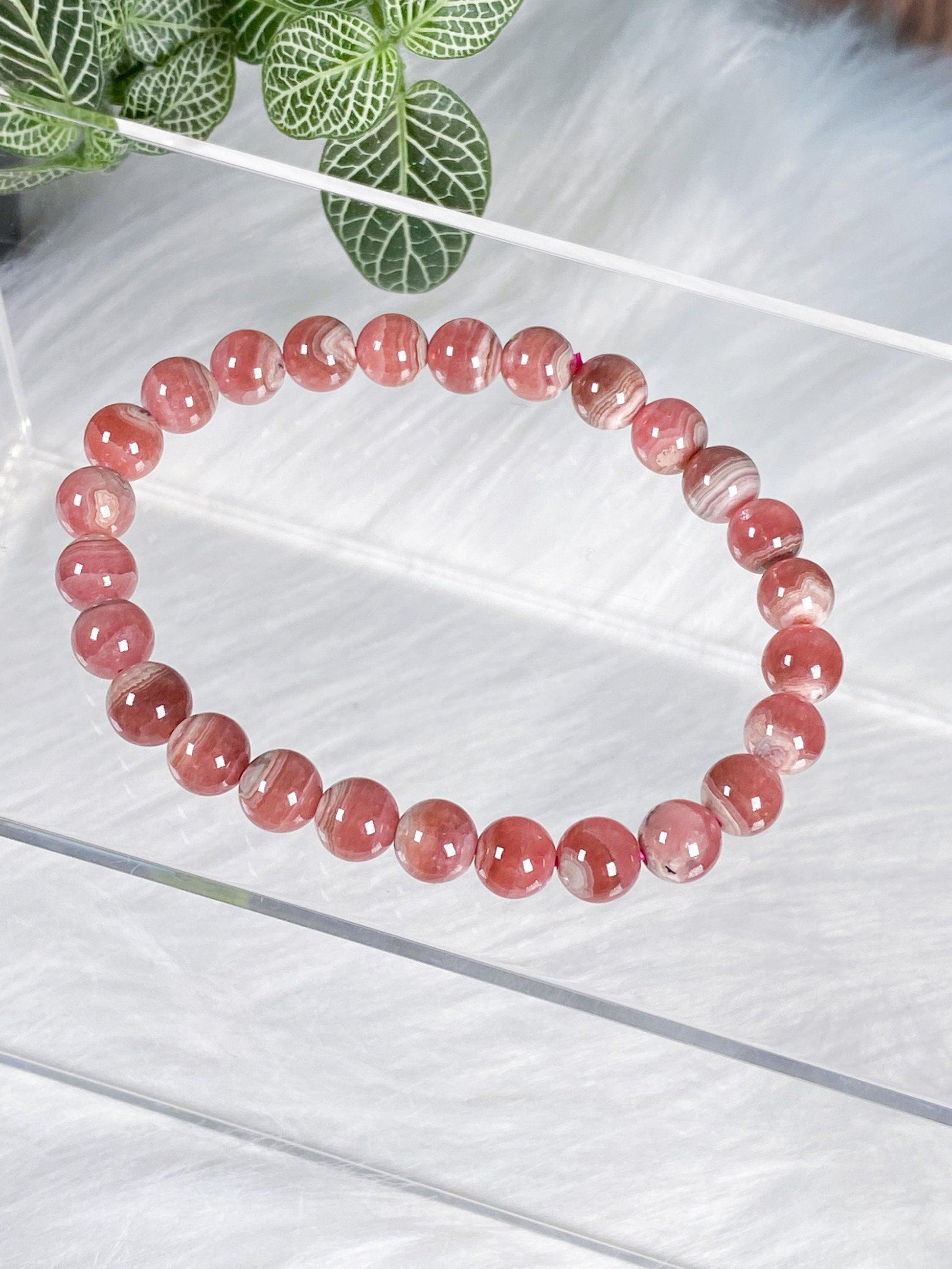 A pink beaded Rhodochrosite Crystal Bracelet from The Crystalary, featuring polished round stones, is elegantly displayed on a clear acrylic stand. In the background, green and white variegated leaves rest on a fuzzy white surface, evoking feelings of self-love.