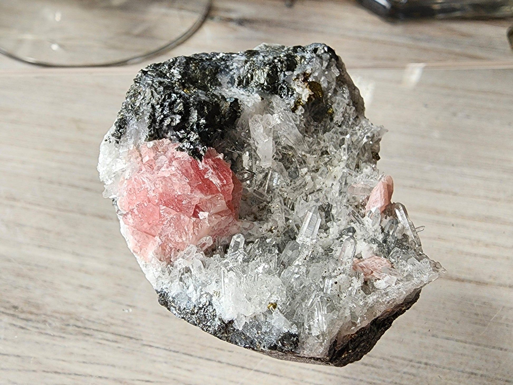 A piece of rough rock from Guangxi, China, embedded with clear Quartz and pink Rhodochrosite crystals from The Crystalary, displayed on a light wooden surface. The sharp, angular needle Quartz crystals contrast strikingly with the dark, rough texture of the rock.