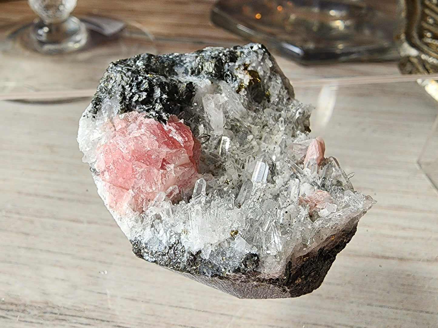 This exquisite Rhodochrosite and Quartz specimen from The Crystalary features translucent needle quartz crystal formations and two prominent clusters of pink rhodochrosite crystals set against a gray, rocky base, displayed on a wooden surface. This remarkable piece hails from the renowned Guangxi region in China.