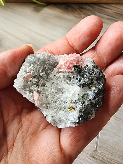 A hand holding a Rhodochrosite, Quartz specimen from Guangxi, China. The clear and white needle Quartz structures are jagged, with small patches of pinkish Rhodochrosite and darker grey and black areas. The background is a wooden surface. Product by The Crystalary.