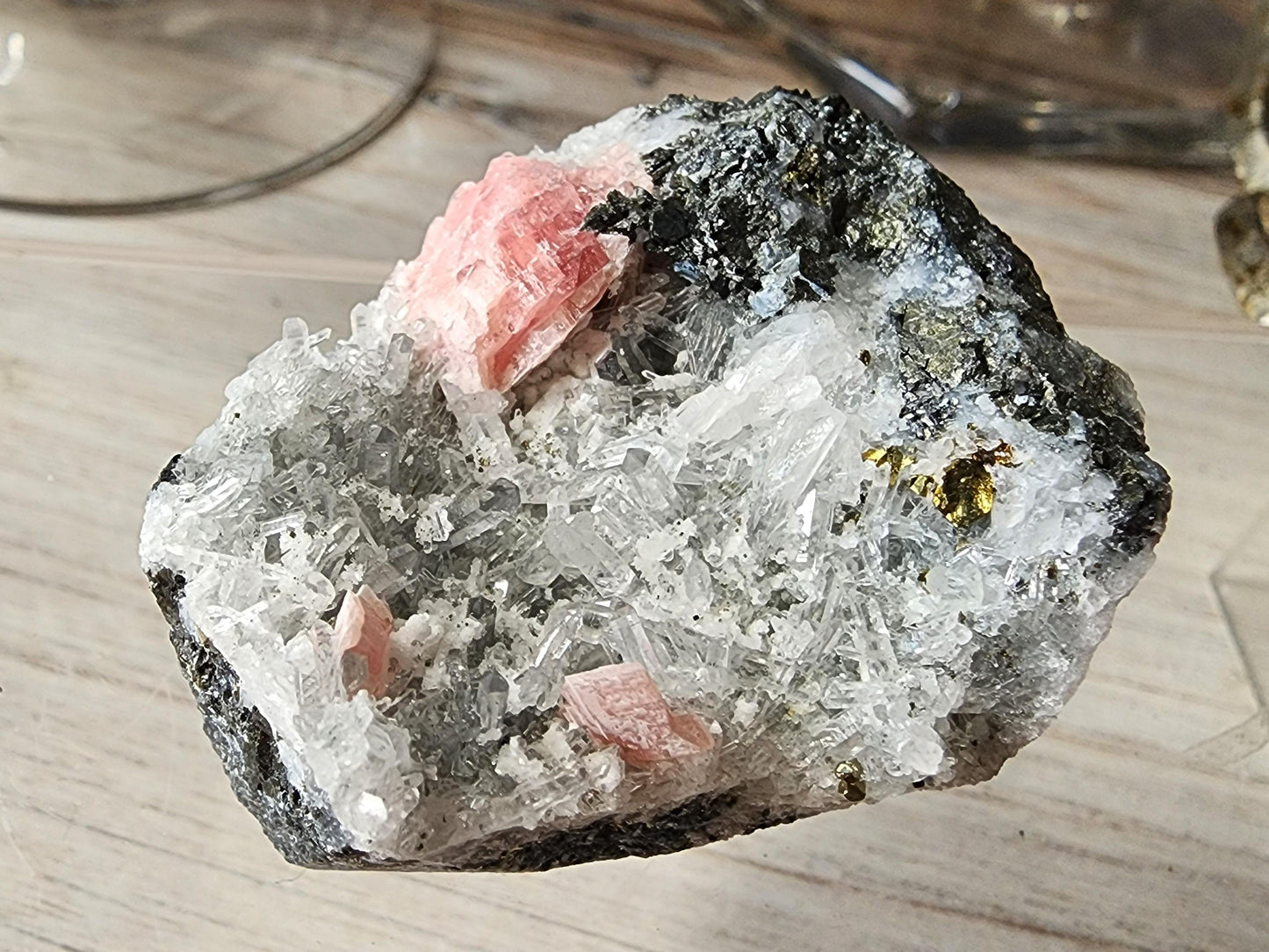 An image of a mineral specimen with clear, crystalline formations and interspersed pink and black sections, featuring needle Quartz from Guangxi. The textured and detailed surface of the Rhodochrosite within the Rhodochrosite, Quartz from Guangxi, China by The Crystalary is highlighted against a wooden background.