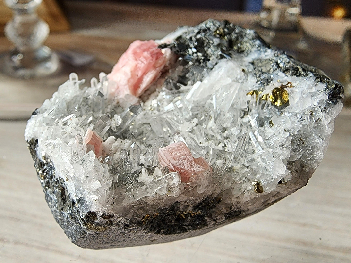 A detailed view of The Crystalary's Rhodochrosite and Quartz specimen from Guangxi, China reveals striking needle-like Quartz formations interlaced with pinkish Rhodochrosite veins within a rugged matrix. The surface sparkles with fine, sharp crystals, accented by subtle metallic gold flecks against the dark background of the stone.