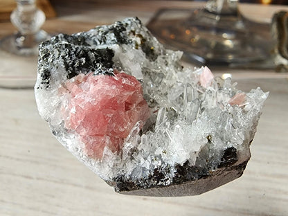 Featured in the collection from The Crystalary, this Rhodochrosite, Quartz specimen from Guangxi, China showcases a prominent pink rhodochrosite formation gracefully embedded in a matrix of clear needle quartz crystals and dark rock-like material. Presented on a light wooden surface with blurred objects in the background, it makes an impressive display piece.