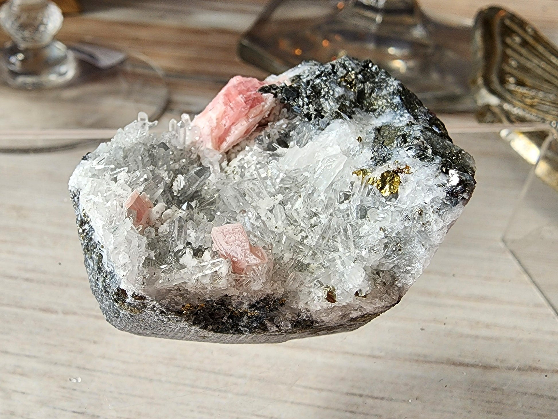 A Rhodochrosite and Quartz mineral specimen from Guangxi, China, offered by The Crystalary, showcasing a blend of clear, white, and pink crystals set in a rough gray stone matrix. The standout pink crystals of Rhodochrosite are beautifully displayed on a wooden surface with a blurred background.