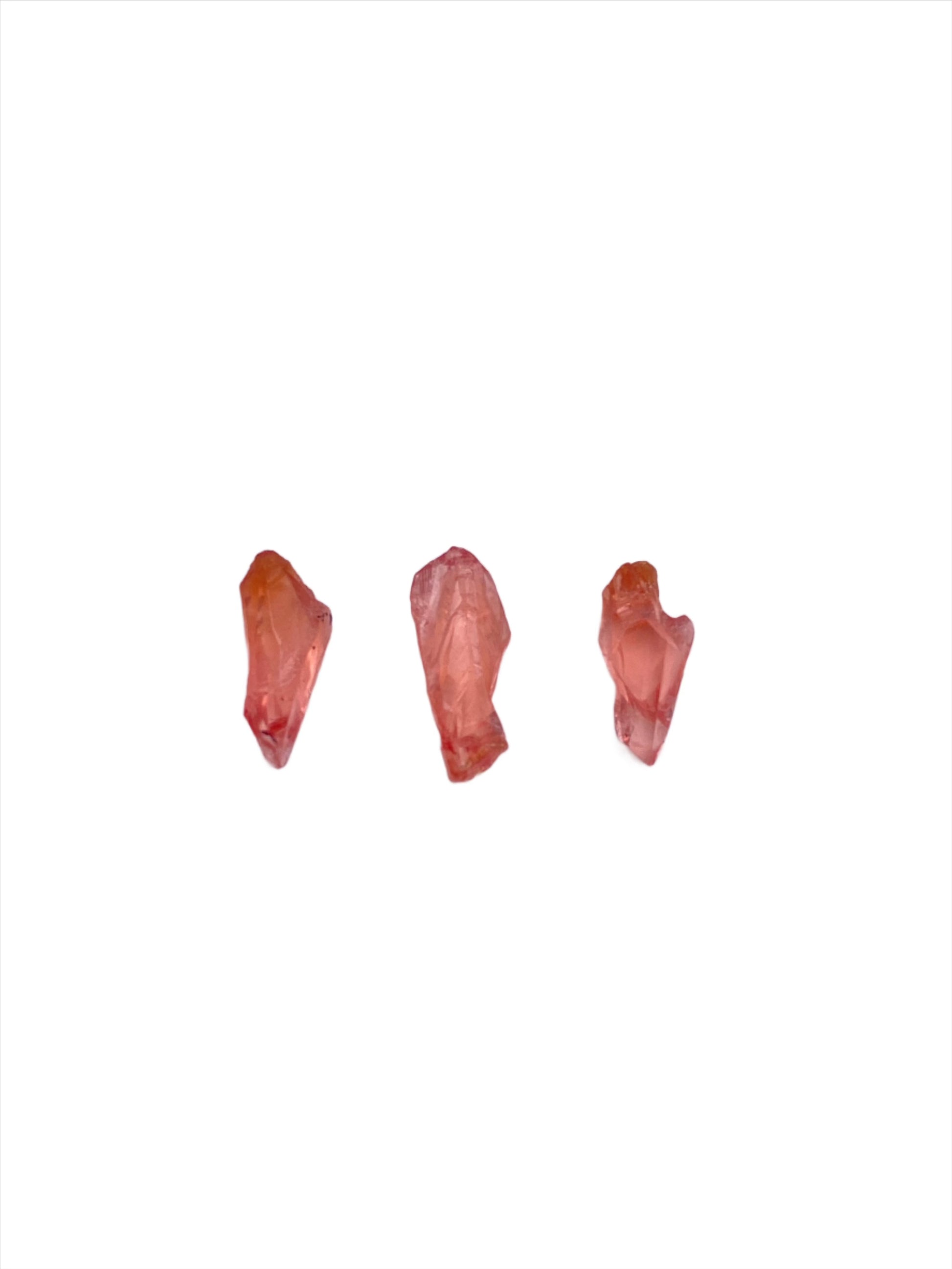 Three small, irregularly shaped Rhodochrosite- Uchucchacua Mine, Lima, Peru crystals from The Crystalary are positioned in a row against a plain white background. The stones vary slightly in shape and size, exhibiting translucent qualities and subtle variations in color, reminiscent of the treasures from the Uchucchacua Mine.