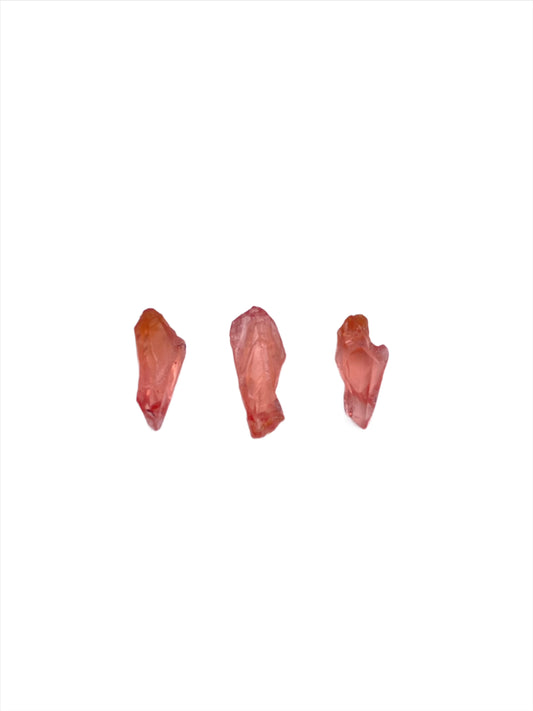 Three small, irregularly shaped Rhodochrosite- Uchucchacua Mine, Lima, Peru crystals from The Crystalary are positioned in a row against a plain white background. The stones vary slightly in shape and size, exhibiting translucent qualities and subtle variations in color, reminiscent of the treasures from the Uchucchacua Mine.