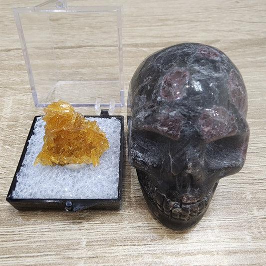 On a wooden surface, a translucent crystal from The Crystalary's collection gleams in an open box next to a polished dark stone skull.