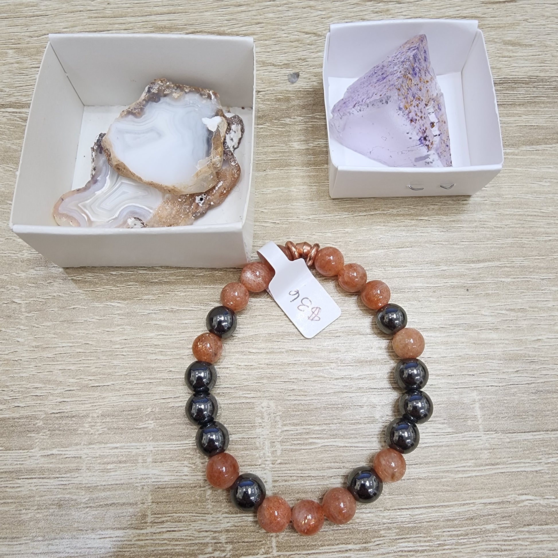 Resting on a wooden surface is a beaded bracelet featuring brown and black beads. Positioned above are two open boxes displaying a slice of agate alongside a shimmering purple crystal. The bracelet, crafted by The Crystalary, comes with an attached small tag displaying the price for "LIVE- sinfullilblonde- 10/3/2024.