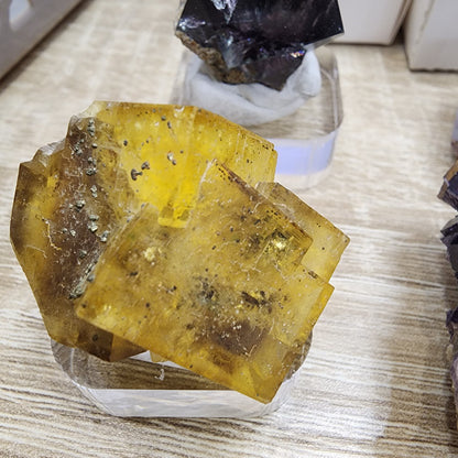 Close-up of a display featuring The Crystalary's product, "LIVE- sinfullilblonde- 10/3/2024," showcasing a large transparent yellow calcite mineral specimen with visible internal patterns. It is elegantly set on a clear stand, resting on a light wood surface and surrounded by other minerals, including an agate and an enchanting sunstone bracelet.