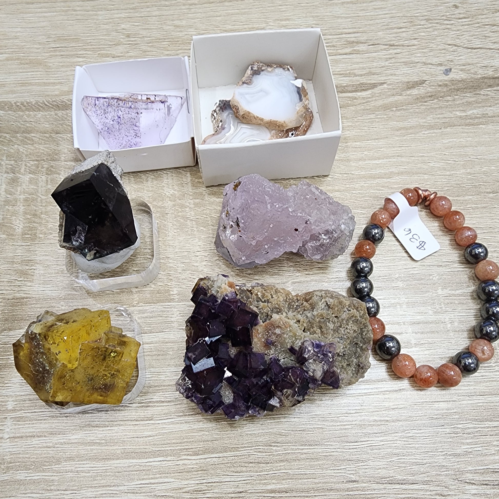 Displayed on a wooden surface is the LIVE-sinfullilblonde-10/3/2024 collection by The Crystalary, featuring an assortment of mineral samples such as agate and fluorite in various shapes and colors—including purple, yellow, and white. Also featured is a sunstone bracelet showcasing mixed beads in brown and black tones with an attached price tag.