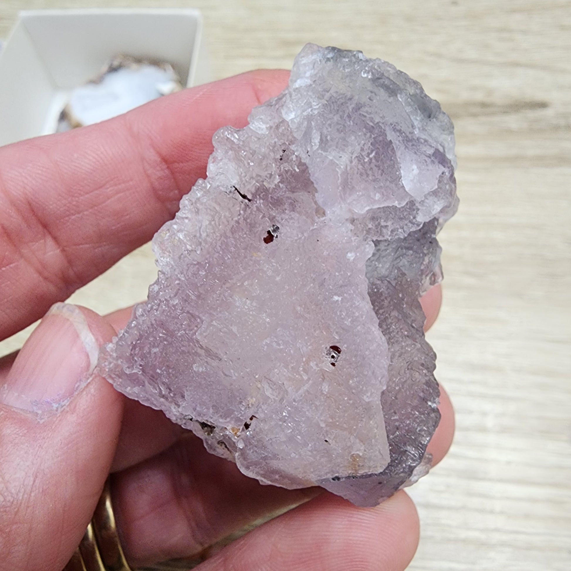 A hand holding The Crystalary's LIVE- sinfullilblonde- 10/3/2024, a rough, translucent pinkish-purple fluorite crystal, against a wooden background.