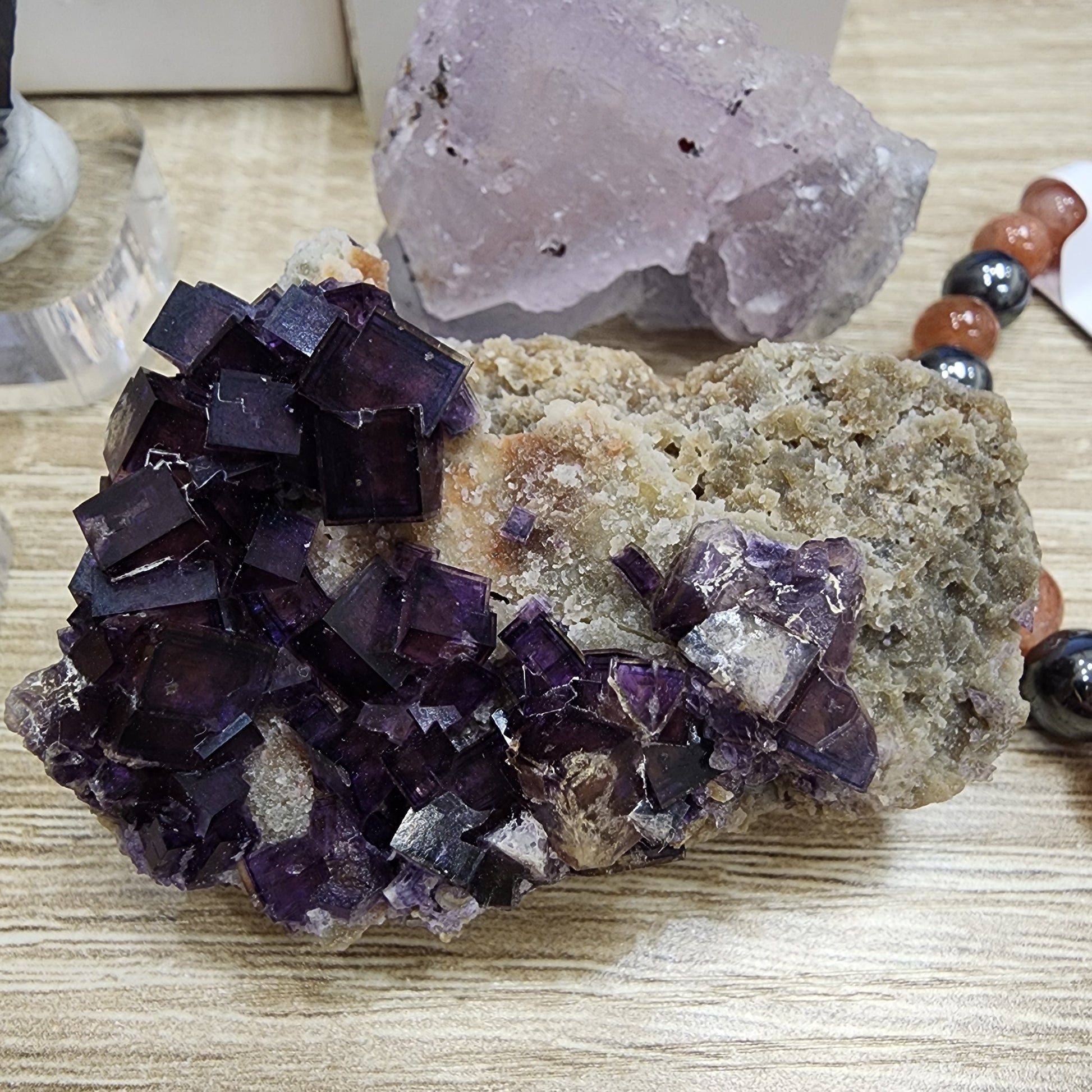 A group of dark purple, square-shaped fluorite crystals is displayed on a rough rock placed on a wooden surface. In the backdrop, a pinkish translucent crystal enhances the beauty of a sunstone bracelet embellished with brown and black beads from The Crystalary's LIVE- sinfullilblonde- 10/3/2024 collection.