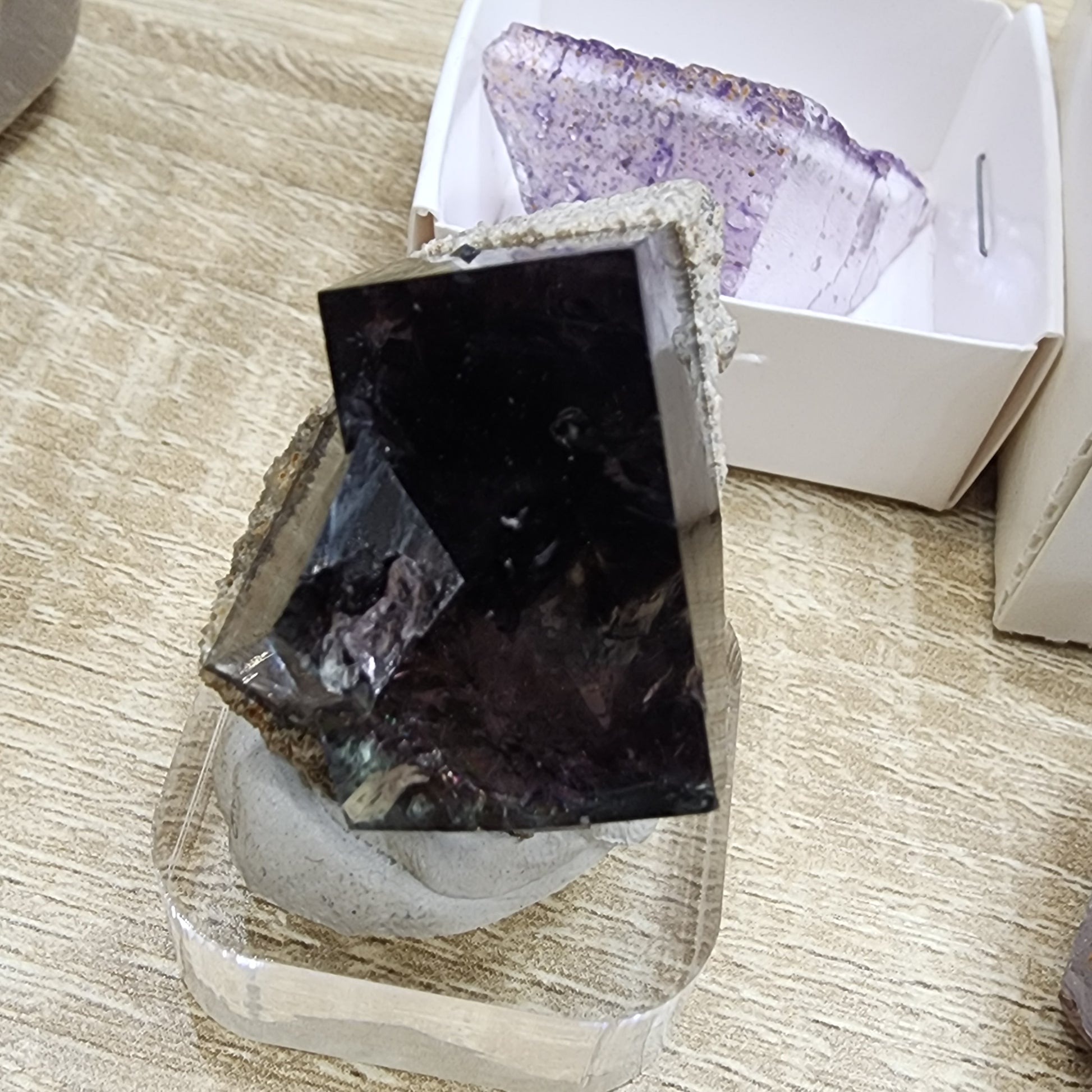 A dark, polished agate crystal from The Crystalary sits elegantly on a stand, with a hint of purple fluorite visible in the background. Both crystals are positioned on a wooden surface, accompanied by nearby white boxes under the LIVE- sinfullilblonde- 10/3/2024 collection.