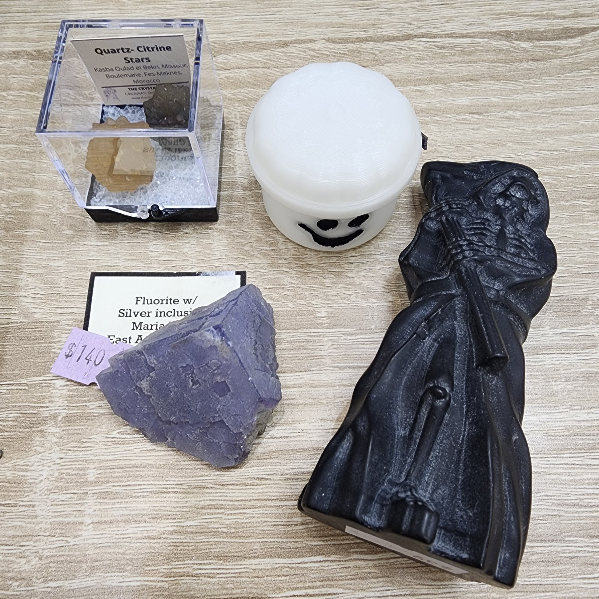 A collection of items on a wooden surface from The Crystalary features a black Grim Reaper carving, a purple fluorite crystal with silver inclusions tagged with the product name LIVE- KarenWebdellRogala- 10/3/2024, a box containing two quartz citrine stars, and a small white container adorned with a smiling face.