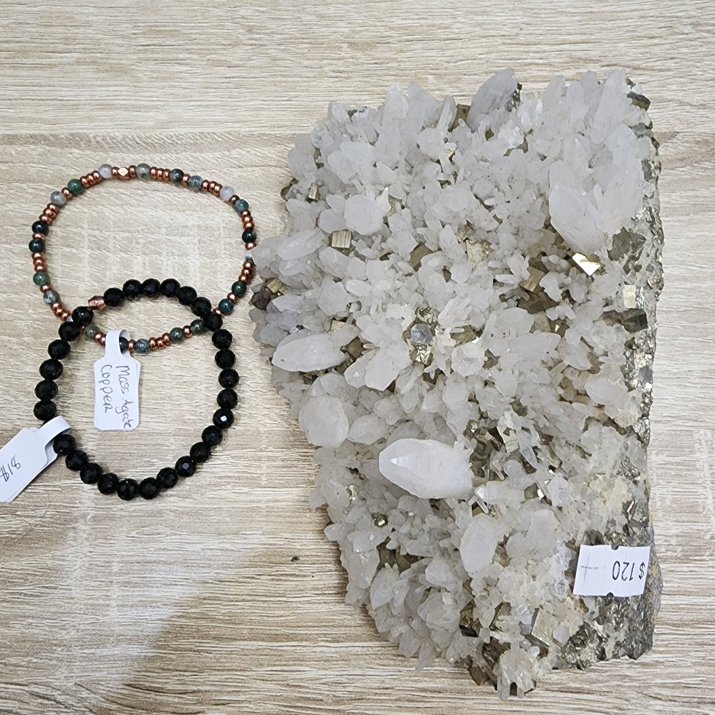 A large cluster of clear quartz crystals, available for $120, is displayed on a wooden surface. Next to it are two beaded bracelets: one featuring green and brown beads with a hint of moss agate priced at $16, and the other adorned with sleek black beads available for $12, all from The Crystalary collection.