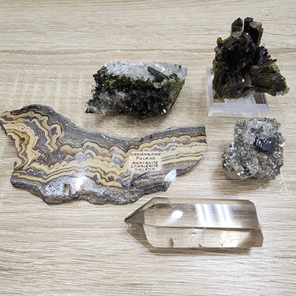 On a wooden surface, The Crystalary from the LIVE- KarenUllrich- 10/3/2024 collection showcases an array of minerals, including a polished slice of banded malachite and a clear quartz crystal point. Among the displayed specimens are an Epidote Duo alongside unpolished treasures with labels, all included in an exciting Quartz Giveaway.