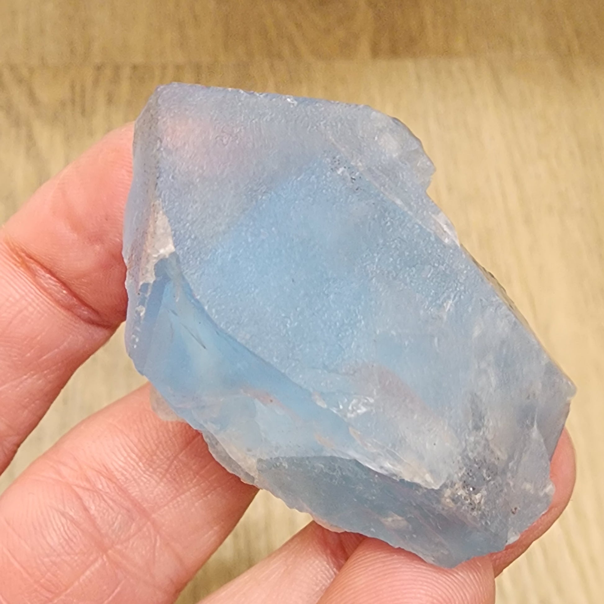 A person's fingers holding a 85-gram, translucent blue and white fluorite crystal with rough and jagged edges from The Crystalary's LIVE- Karenwebdellrogala- 9/27/24 collection, against a light brown wooden background.