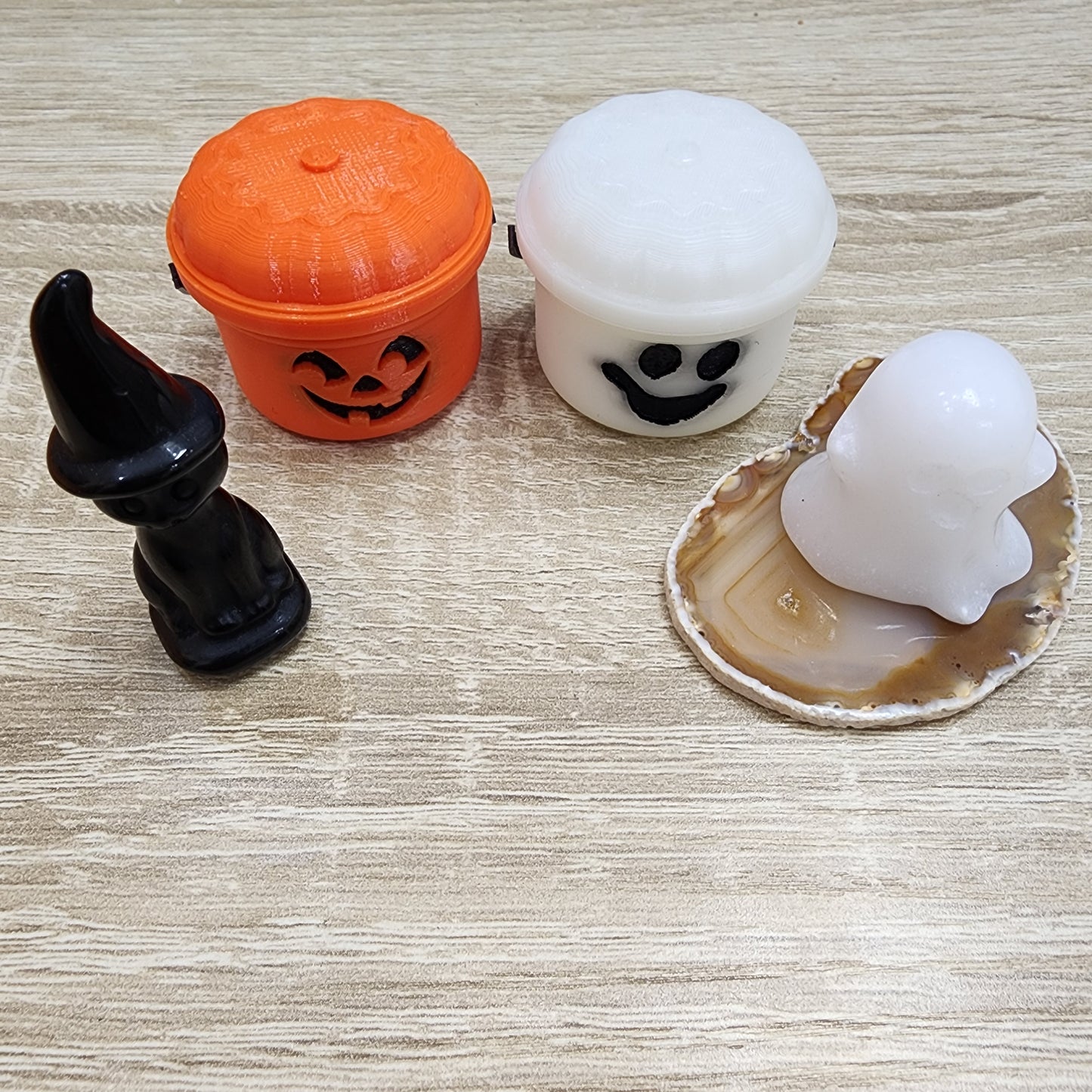 A collection of Halloween-themed pieces from The Crystalary, featuring a black witch figurine, an orange jack-o'-lantern container, and a white ghost container, are arranged on a wooden surface. Accompanying these is a white ghost figurine elegantly perched on an agate slice. Adding to the mysterious ambiance of this eerie ensemble is the presence of a LIVE-LewisThomas-10/3/2024 carved obsidian cat.