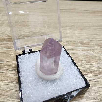 Displayed elegantly on a wooden surface, the LIVE- PrincessPelvis- 11/14/24 from The Crystalary is a captivating purple crystal that resembles a vibrant purpurite. It rests on a white base within a sleek black and clear plastic case, beautifully capturing the light with its reflective, faceted design.