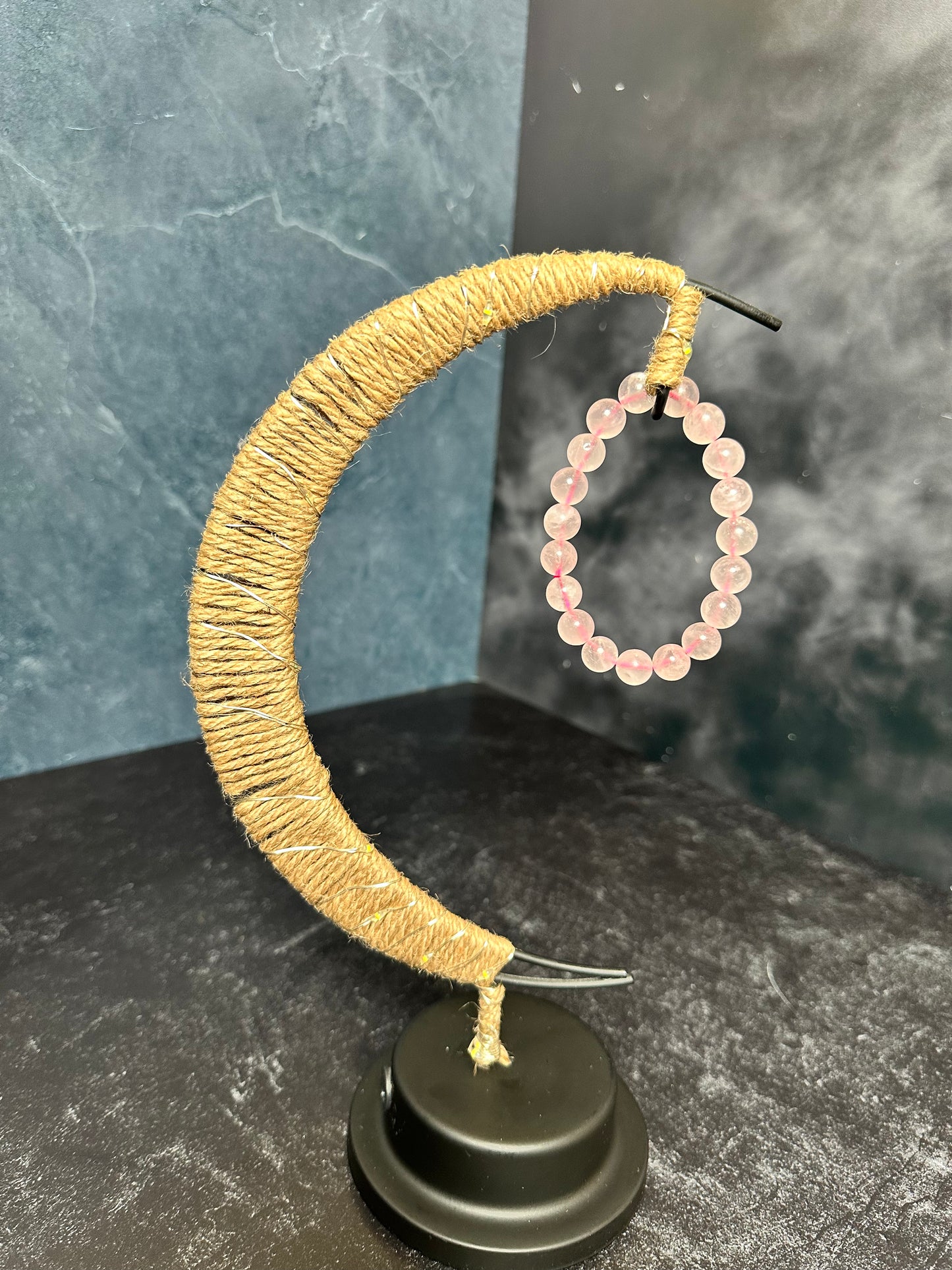 A handmade moon-shaped jewelry holder wrapped in twine, displaying The Crystalary's Rose Quartz Crystal Bracelet hanging from its curved end. The background features a dark-tiled surface with a smoky texture, perfectly complementing the elegance of ethical crystal and gem trade pieces.