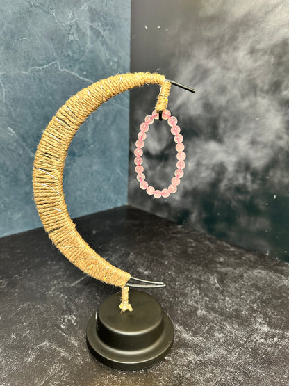 A handmade crescent moon stand wrapped in rustic twine holds The Crystalary's Rose Quartz Crystal Bracelet. The stand features a black base and is set against a dark, textured background, highlighting the bracelet's individual beads sourced from the ethical crystal and gem trade.