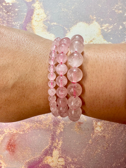 A close-up of a wrist adorned with three exquisite Rose Quartz Crystal Bracelets by The Crystalary, each made of round, translucent pink beads. The background features a textured surface with pink and gold hues. The hand is slightly tanned, showcasing the beauty and ethics of The Crystalary's gemstone bracelets trade.