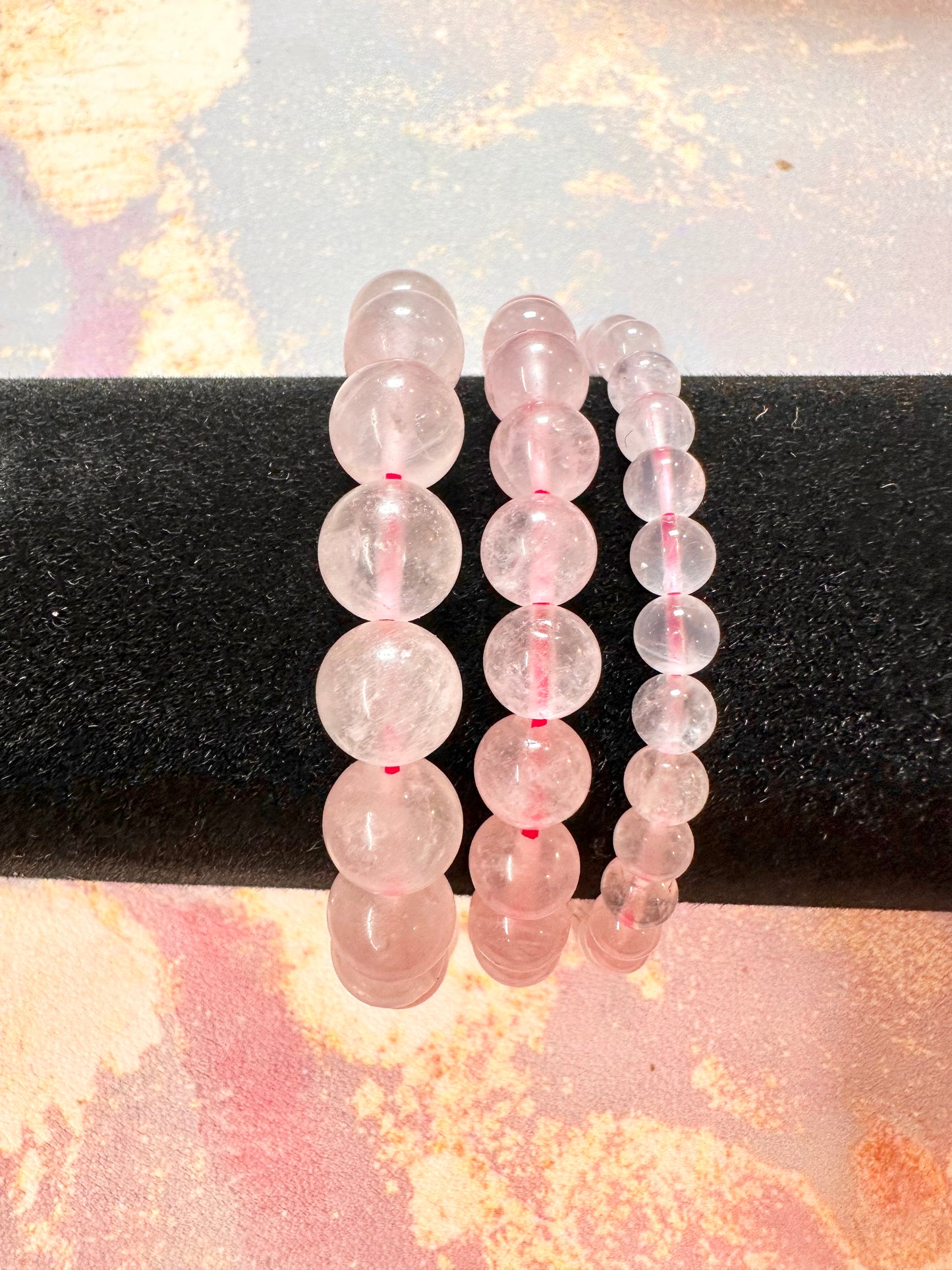 Three Rose Quartz Crystal Bracelets by The Crystalary are elegantly displayed on a black cylindrical stand, showcasing beads with sizes ranging from small to large. The background features a colorful, abstract design with pink, yellow, and blue tones, highlighting the sophistication of these handcrafted gemstone bracelets made from ethically sourced crystals.