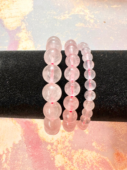 Three Rose Quartz Crystal Bracelets by The Crystalary are elegantly displayed on a black cylindrical stand, showcasing beads with sizes ranging from small to large. The background features a colorful, abstract design with pink, yellow, and blue tones, highlighting the sophistication of these handcrafted gemstone bracelets made from ethically sourced crystals.