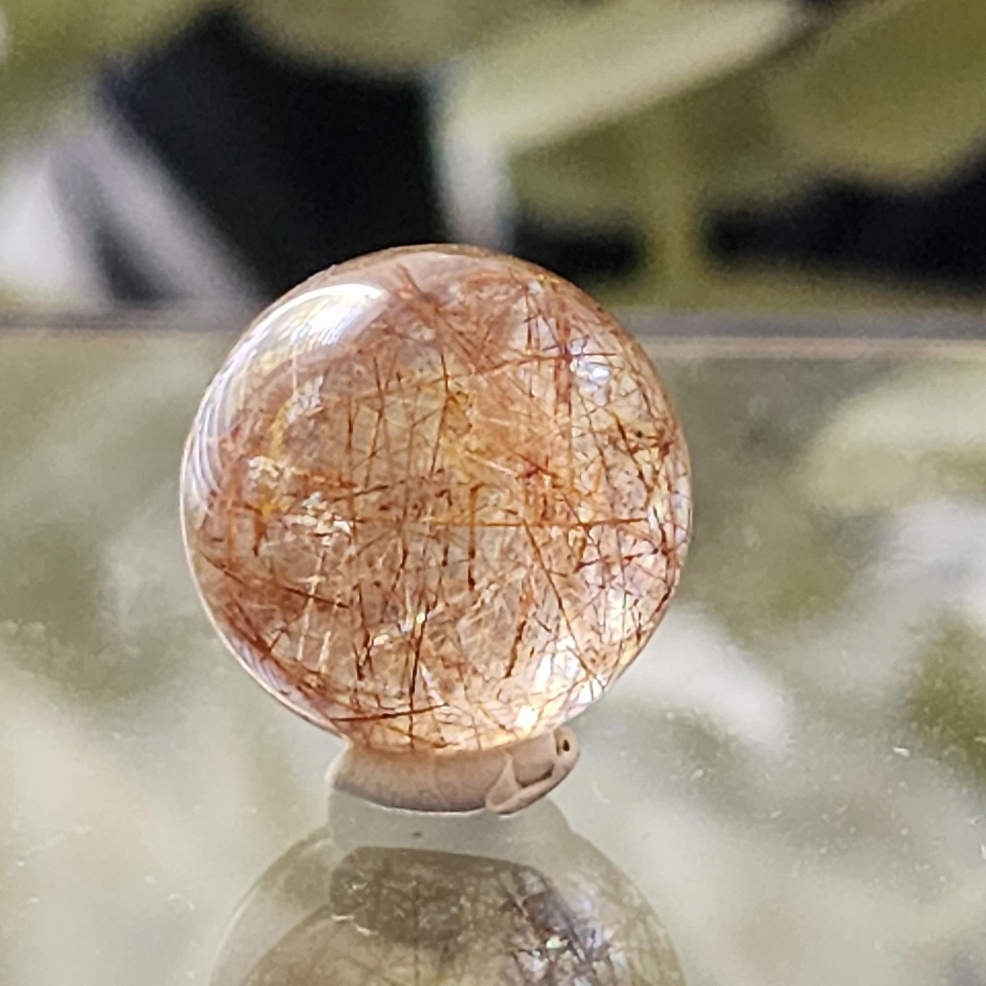 Close-up of the Rutile Quartz mini sphere 13mm from The Crystalary showcasing its intricate golden and copper inclusions, set against a softly blurred background. This polished 13mm gemstone rests on a small stand or putty, reflecting light subtly.