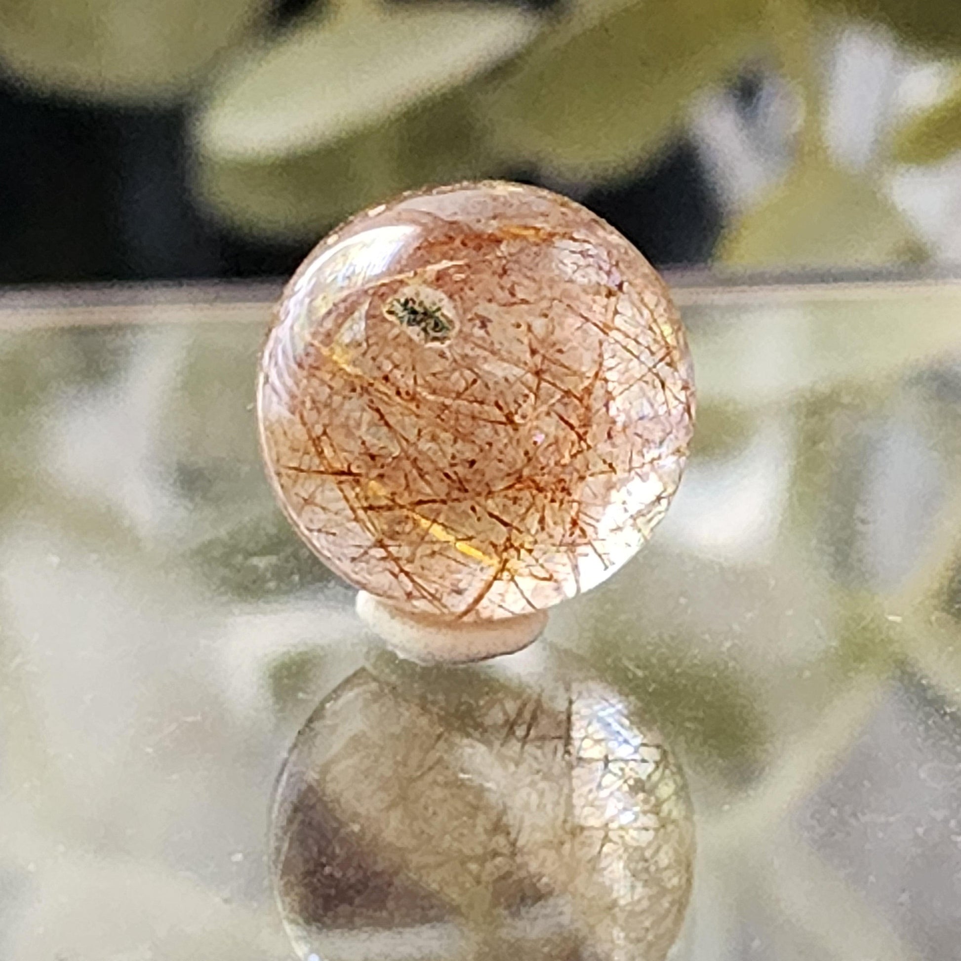 A Rutile Quartz mini sphere 13mm from The Crystalary, with clear quartz and golden needle-like inclusions, rests on a reflective surface. The blurred background hints at green foliage, adding a natural aesthetic. The crystal's internal patterns and reflections create a visually intricate effect.