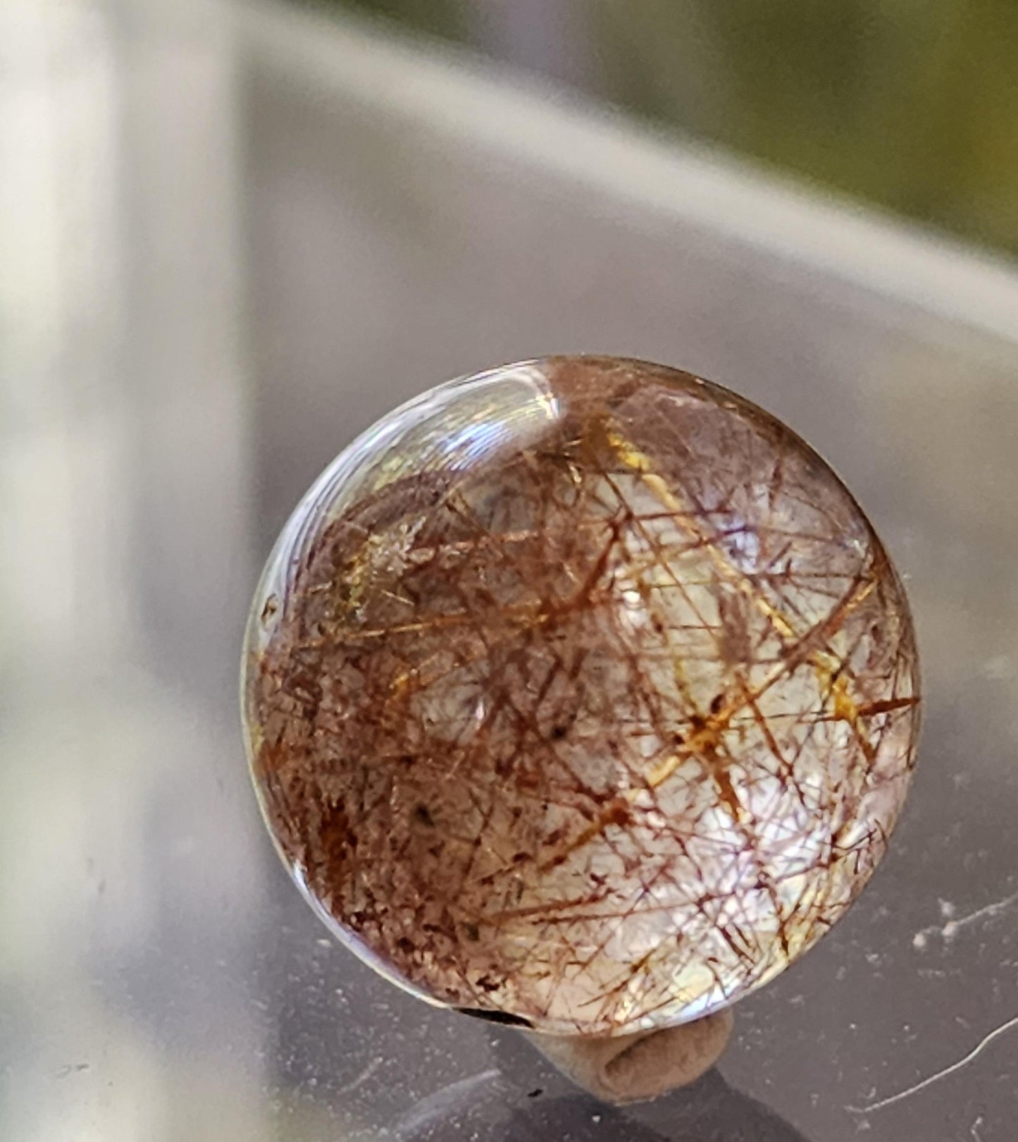 A close-up image showcases The Crystalary's Rutile Quartz mini sphere (13mm), a clear, spherical orb featuring delicate, brownish-gold thread-like inclusions. The polished crystal is elegantly resting on a reflective surface, with the softly blurred background accentuating the orb's intricate details.