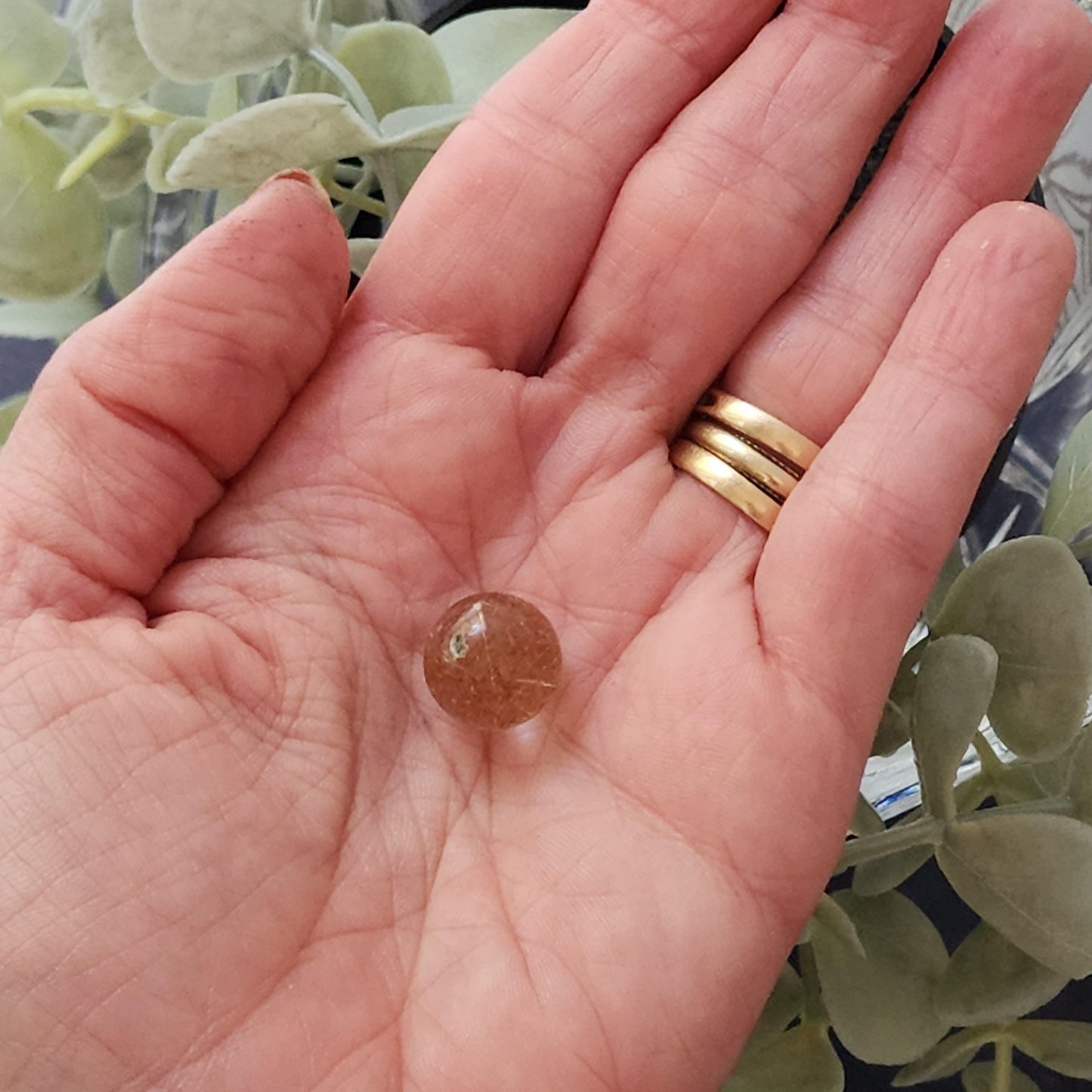 A close-up of a hand holding a small, translucent brown sphere. The hand, adorned with a gold ring, gently cradles what appears to be The Crystalary's Rutile Quartz mini sphere (13mm) against a blurred background of green leaves.