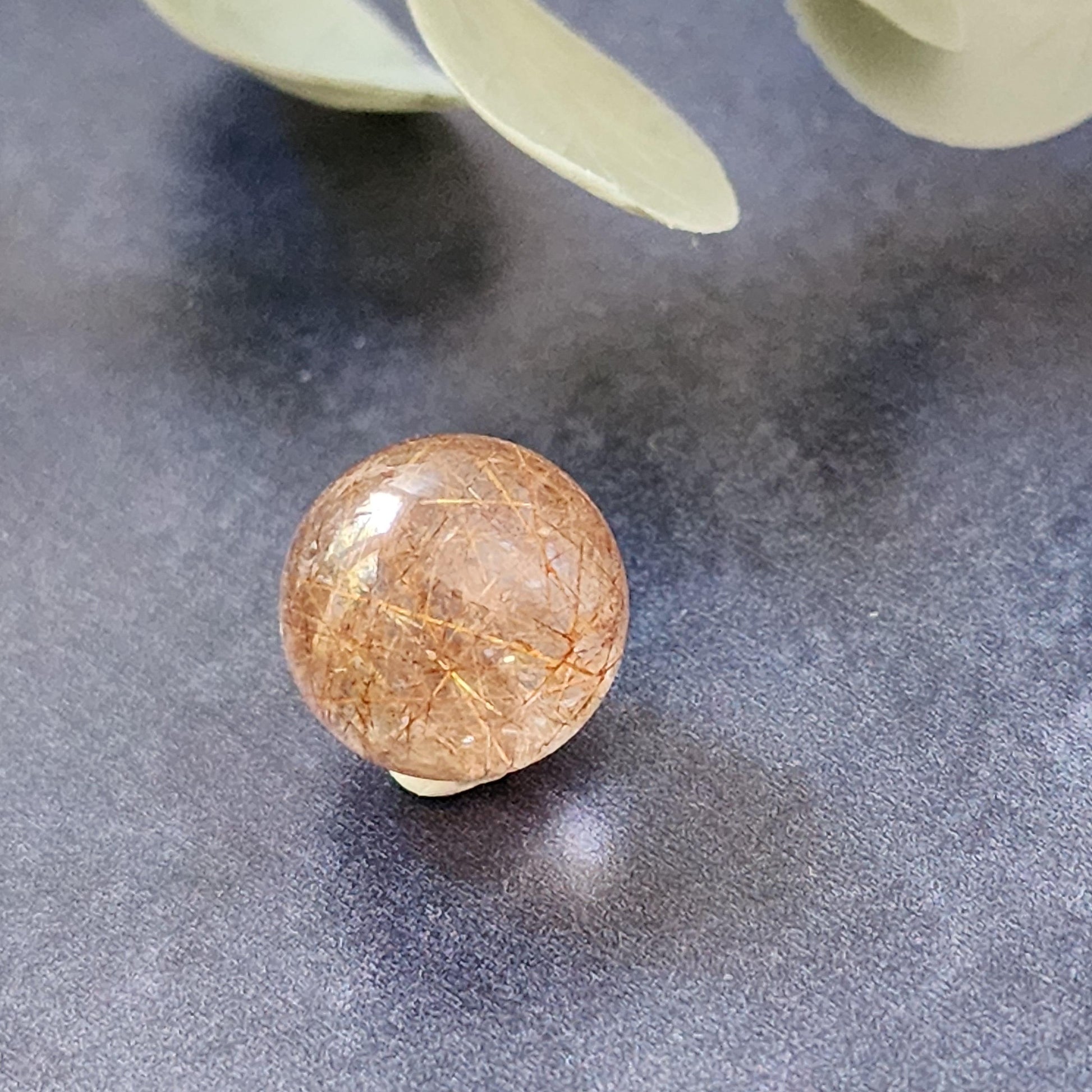 A round, semi-transparent Rutile Quartz mini sphere from The Crystalary, measuring 13mm and featuring visible needle-like inclusions, is placed on a dark, textured surface. Some green leaves are slightly visible in the background.