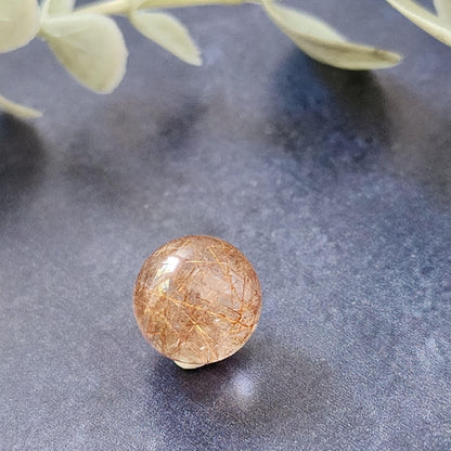 A 13mm Rutile Quartz mini sphere from The Crystalary rests on a dark, textured surface. The polished crystal displays a transparent base with delicate, golden needle-like inclusions inside. Out-of-focus green leaves are visible in the background.