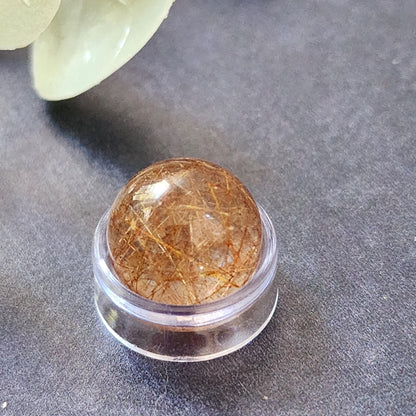 A close-up photo of a Rutile Quartz mini sphere 13mm from The Crystalary, showcasing its round, transparent form with reddish-brown needle-like inclusions. The polished gemstone is elegantly displayed on a small, round, clear plastic stand against a dark, textured background. This piece is perfect for collectors or as worry stones to ease anxiety.