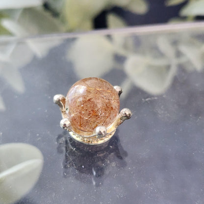 Close-up of a polished Rutile Quartz mini sphere 13mm by The Crystalary, set in a simple, elegant metallic stand with prongs. The quartz features visible needle-like inclusions and rests on a reflective surface. Blurred green foliage in the background evokes the classic soothing quality of worry stones.