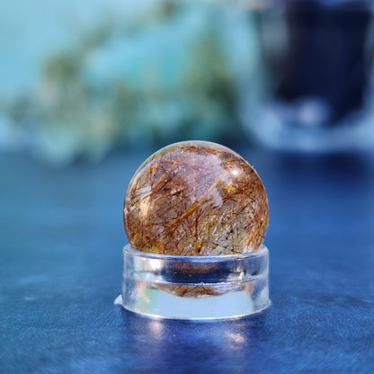 A Rutile Quartz mini sphere from The Crystalary, with a diameter of 15mm, showcases golden needle-like inclusions on a clear stand against a blurred blue-green background. The polished quartz sphere exhibits an intricate web of fine, shimmering lines within its transparent body.