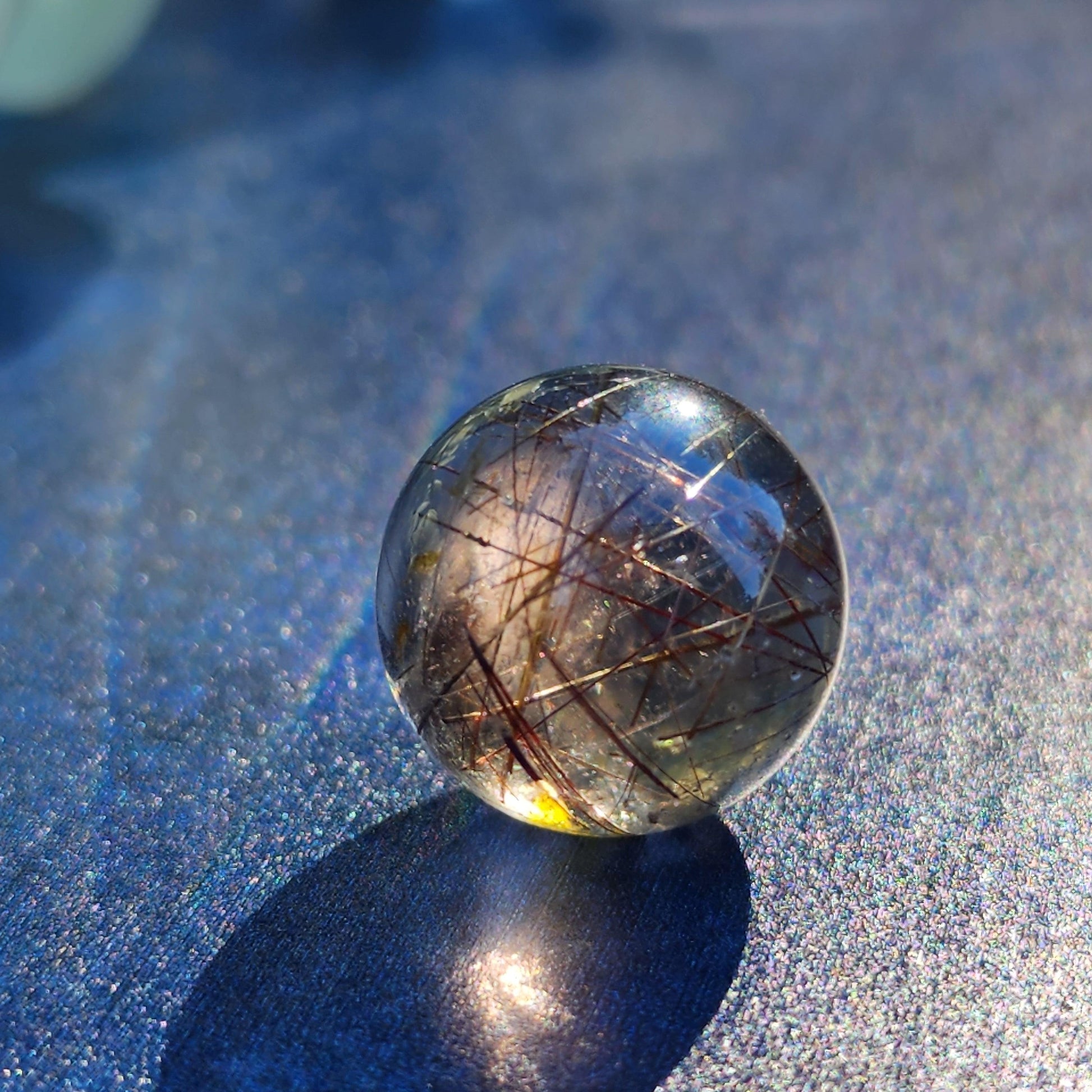 A close-up of The Crystalary's Rutile Quartz mini sphere (15mm) with embedded hair-like inclusions stands on a glittering blue surface. The sunlight casts a shadow that highlights the iridescent qualities of both the mini sphere and the background, reminiscent of Rutile Quartz used in meditation practices.