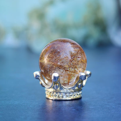 A polished Rutile Quartz mini sphere 15mm from The Crystalary, featuring streaks of brown and gold, is placed on a small silver crown. The background is softly blurred, highlighting the detailed texture and reflections on both the gemstone and crown.
