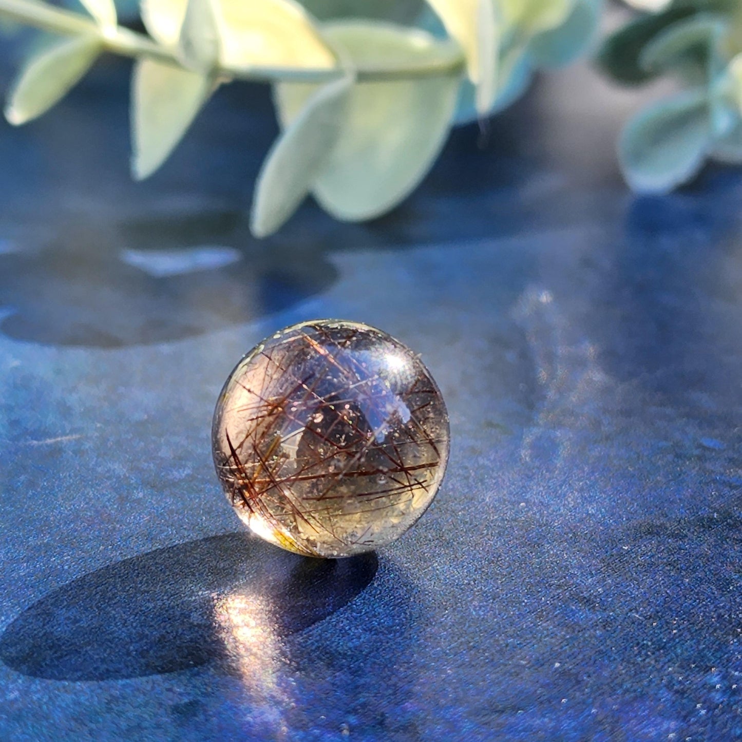 A close-up photograph of The Crystalary's Rutile Quartz mini sphere (15mm) resting on a blue surface. The clear sphere contains fine strands of black and gold, creating a captivating, intricate pattern. Soft green leaves are blurred in the background, adding a natural element to the image's serene meditation setting.