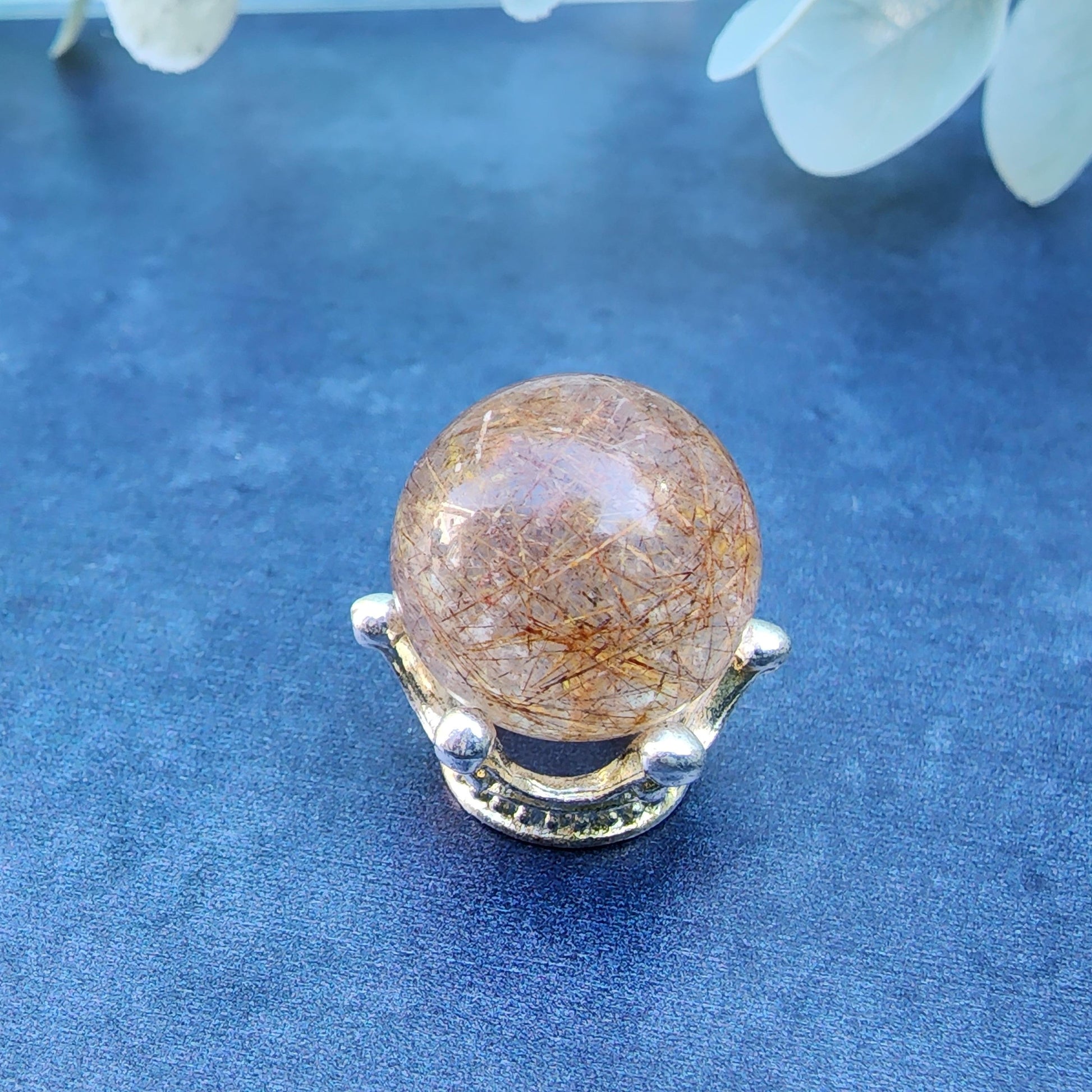 Displayed on a decorative silver stand against a dark blue background, the Rutile Quartz mini sphere (15mm) from The Crystalary showcases its transparent golden hue. Inside the polished crystal, delicate golden needle-like inclusions of Rutile Quartz are visible. Eucalyptus leaves are partially visible in the upper right corner.