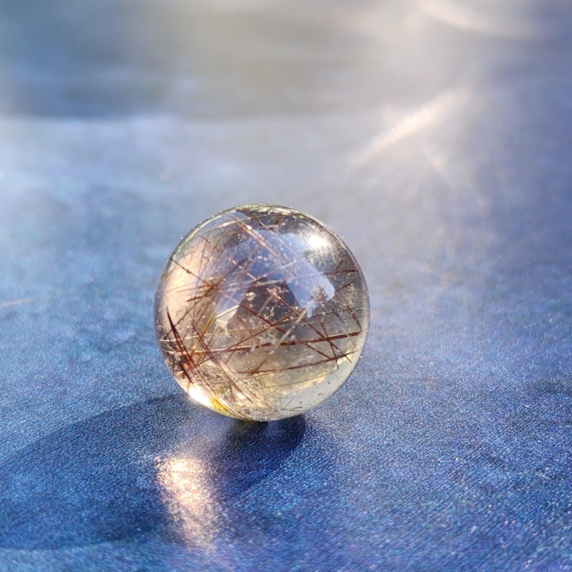 A Rutile Quartz mini sphere from The Crystalary, measuring 15mm, rests on a smooth, reflective blue surface. Intricate brownish-gold inclusions inside the clear sphere catch the light, enhancing its luminous appearance and giving it a radiant, ethereal quality perfect for meditation.