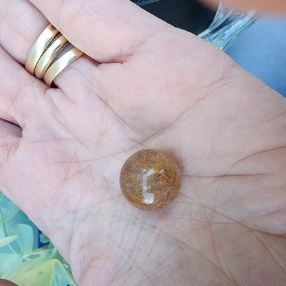 A hand holding a Rutile Quartz mini sphere (15mm) from The Crystalary, showcasing its polished surface with brown and golden hues. The person's fingers are adorned with several gold rings. The background is slightly blurry, featuring hints of blue and green.