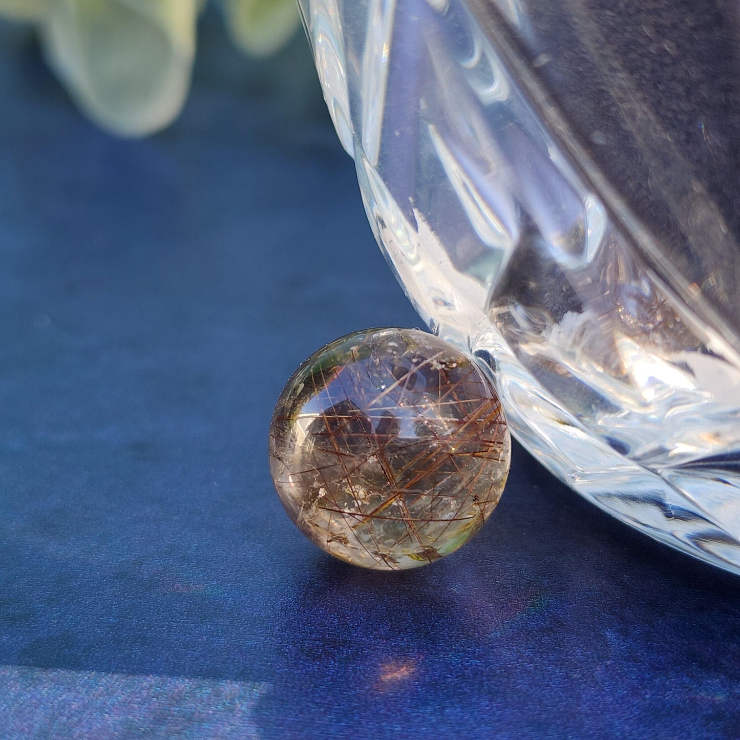 A rounded, translucent Rutile Quartz mini sphere 15mm from The Crystalary with delicate golden threads sits on a reflective blue surface next to a clear, faceted glass object, creating an elegant arrangement perfect for meditation, with soft focus greenery in the background.