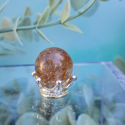 A round, amber-colored Rutile Quartz mini sphere 15mm from The Crystalary sits atop a silver crown-shaped pedestal. The crown has small silver spheres and intricate detailing. Soft-focus green leaves in the background create a serene, natural ambiance that is beautifully reflected on a mirrored surface.